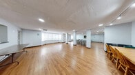 Located at 545-565 Belmont Ave W in Kitchener, Ontario, this single-floor multipurpose space offers a generous and versatile layout well-suited for gatherings, meetings, or community activities. The main open area is characterized by warm hardwood-style flooring that extends throughout, complemented by light grey walls and a textured ceiling with recessed and track lighting that ensures bright, even illumination across the space.

The primary gathering area is expansive, accommodating multiple folding table-and-chair configurations, making it highly adaptable for various uses. A large bay window along one wall features roller blinds and integrated baseboard heating beneath, allowing for natural light while maintaining comfort. Decorative artwork is displayed on the walls, adding a welcoming character to the otherwise functional setting.

A built-in bookshelf unit adds both practical storage and visual interest to one section of the space. The layout also includes a separate kitchen or servery area finished with tile flooring, cabinetry, and a counter — ideal for food preparation and service during events.

Additional features include clearly marked emergency exits, fire safety equipment, and interior glass-paneled doors that connect distinct zones within the space. The overall design is clean, practical, and well-maintained, providing a functional environment suitable for a wide range of community or organizational purposes.