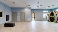 This well-appointed amenity facility spans a single floor and is thoughtfully designed to support both fitness and community activities. The exercise room is the standout feature, offering an expansive, light-filled space outfitted with a comprehensive array of equipment, including treadmills, cable machines, stationary bikes, free weights, and specialty strength machines. A striking, large-scale abstract mural anchors one wall of the main fitness area, adding a bold artistic element to the space. Patterned, textured carpet tiles in muted green and teal tones cover the floors throughout the gym, providing comfort underfoot. Multiple windows line the perimeter, inviting natural light and views of the surrounding exterior. A dedicated secondary fitness alcove features a wall-mounted television and additional equipment, ideal for focused training. The living room area presents a clean, open layout with soft blue-gray walls and an intricate geometric parquet-style floor, creating a calm and flexible environment suitable for group gatherings or events. Linear LED lighting runs across the ceilings in both areas, ensuring consistent, modern illumination throughout. Multiple access doors connect the spaces seamlessly.