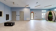 This well-appointed amenity facility spans a single floor and is thoughtfully designed to support both fitness and community activities. The exercise room is the standout feature, offering an expansive, light-filled space outfitted with a comprehensive array of equipment, including treadmills, cable machines, stationary bikes, free weights, and specialty strength machines. A striking, large-scale abstract mural anchors one wall of the main fitness area, adding a bold artistic element to the space. Patterned, textured carpet tiles in muted green and teal tones cover the floors throughout the gym, providing comfort underfoot. Multiple windows line the perimeter, inviting natural light and views of the surrounding exterior. A dedicated secondary fitness alcove features a wall-mounted television and additional equipment, ideal for focused training. The living room area presents a clean, open layout with soft blue-gray walls and an intricate geometric parquet-style floor, creating a calm and flexible environment suitable for group gatherings or events. Linear LED lighting runs across the ceilings in both areas, ensuring consistent, modern illumination throughout. Multiple access doors connect the spaces seamlessly.