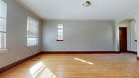 This single-floor space showcases a thoughtful blend of classic architectural character and practical modern updates. Throughout the unit, warm hardwood floors and rich dark wood baseboards and door trim create a cohesive, timeless aesthetic complemented by soft gray-toned walls and crisp crown molding along the ceilings.

A standout feature is the graceful arched doorway that connects the main living area to an inner hallway, adding architectural elegance to the layout. The living spaces are well-lit by multiple windows that invite natural light, and a wall-mounted ductless mini-split system provides efficient climate control. Substantial panel doors with period-appropriate hardware reinforce the traditional character of the space.

The kitchen is equipped with cream-colored shaker-style cabinetry featuring dark iron hardware, granite-look countertops, a beadboard backsplash, and a window above the sink for natural light. The layout offers generous counter and storage space.

The bathroom is finished with classic white subway tile walls and a black-and-white basketweave tile floor — a timeless combination that lends a clean, vintage-inspired look. A hallway connects the bathroom to the rest of the unit, with dark wood doors visible throughout.

Additional rooms include generously sized bedrooms with built-in closets, hardwood floors, and ample window placement for natural light, completing a well-rounded and characterful living space.