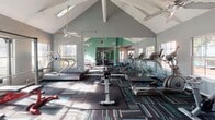 This fitness center in Gainesville, Florida, offers a spacious, single-floor layout designed to accommodate a comprehensive workout experience. The facility's most striking architectural feature is its soaring vaulted ceiling with exposed white beams, which creates an open, airy atmosphere throughout the main exercise area. Ceiling fans are positioned throughout to promote air circulation, complementing the natural light that pours in through multiple windows lining the walls.

The flooring consists of a distinctive patterned carpet in teal, blue, and gray tones, with rubber mat sections providing traction in key workout zones. Full-length mirrors are mounted along multiple walls, visually expanding the space and allowing users to monitor their form during exercise.

The cardio zone is well-appointed with multiple treadmills, elliptical trainers, and stationary bikes. A connected strength-training area features a cable machine, a dumbbell rack with a range of free weights, a punching bag, and adjustable benches for resistance exercises. The teal-painted accent walls carry consistently throughout both sections, unifying the space with a cohesive, energetic color palette that enhances the overall workout environment.