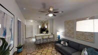 This single-floor space showcases an open-concept layout that seamlessly connects the living area and kitchen, creating a cohesive and functional environment. The living area is anchored by a contemporary gray sofa positioned atop a geometric-patterned area rug, complemented by a decorative wall panel featuring a honeycomb design. A ceiling fan with an integrated light fixture serves as a central overhead element, providing both airflow and ambient lighting.

The kitchen flows directly from the living space, featuring a peninsula-style breakfast bar with white countertops and bar seating for multiple guests. Pendant lights hang above the bar, adding focused task lighting and a design accent. The kitchen is equipped with dark cabinetry and stainless steel appliances, including a refrigerator. Track lighting runs along the kitchen ceiling for additional illumination.

Wood-look flooring extends throughout the space, adding warmth and visual continuity. White interior doors are visible, including what appears to be a front entry door. The neutral grey-and-white color palette, accented with teal and natural tones, contributes to the space's modern and inviting ambiance.