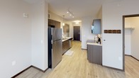 This single-floor unit offers a thoughtfully designed open layout that balances functionality and modern style. Light wood-look luxury vinyl flooring flows consistently throughout the space, creating a cohesive and clean aesthetic.

The kitchen is a standout feature, equipped with dark wood cabinetry, white quartz countertops, a double stainless steel sink, stainless steel appliances including a full-size refrigerator and dishwasher, and a white subway tile backsplash. Pendant lighting and track lighting illuminate the space effectively, while upper cabinetry provides ample storage. The kitchen connects seamlessly to an entry hallway with a dark wood front door.

The living area is generously proportioned, offering an open and versatile space. A ceiling fan with integrated lighting anchors the room, and a window with blinds allows for natural light. A wall-mounted HVAC unit provides individual climate control.

The bathroom is well-sized and features warm tan ceramic tile wainscoting along the lower walls, complemented by light wood-look tile flooring. A long vanity with dark wood cabinetry and a white countertop offers substantial storage and counter space. The overall design palette is neutral and contemporary, with white upper walls and clean finishes throughout, creating a practical and inviting environment.