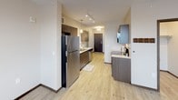 This single-floor unit offers a thoughtfully designed open layout that balances functionality and modern style. Light wood-look luxury vinyl flooring flows consistently throughout the space, creating a cohesive and clean aesthetic.

The kitchen is a standout feature, equipped with dark wood cabinetry, white quartz countertops, a double stainless steel sink, stainless steel appliances including a full-size refrigerator and dishwasher, and a white subway tile backsplash. Pendant lighting and track lighting illuminate the space effectively, while upper cabinetry provides ample storage. The kitchen connects seamlessly to an entry hallway with a dark wood front door.

The living area is generously proportioned, offering an open and versatile space. A ceiling fan with integrated lighting anchors the room, and a window with blinds allows for natural light. A wall-mounted HVAC unit provides individual climate control.

The bathroom is well-sized and features warm tan ceramic tile wainscoting along the lower walls, complemented by light wood-look tile flooring. A long vanity with dark wood cabinetry and a white countertop offers substantial storage and counter space. The overall design palette is neutral and contemporary, with white upper walls and clean finishes throughout, creating a practical and inviting environment.