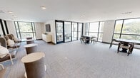 Located at 5102 Station Dr in Birmingham, Alabama, this single-floor amenity space offers a thoughtfully designed environment that blends comfort with contemporary style. The open-plan layout encompasses a combined living and dining area, bathed in abundant natural light courtesy of expansive floor-to-ceiling windows with sleek black frames that offer sweeping views of the surrounding greenery and neighborhood streetscape.

The living area is appointed with high-backed lounge chairs, cylindrical side tables, and a wall-mounted flat-screen television set atop a cream-toned credenza — creating an inviting setting for casual gatherings or relaxed socializing. Adjacent to this lounge zone, a dining area provides multiple seating arrangements with dark wood tables and mixed seating options suited for both collaborative and independent use.

A glass-walled private conference or dining room is accessible via a bold black-framed door, offering a more intimate setting centered around a long white table with upholstered chairs and anchored by a statement pendant light. This enclosed room benefits from its own set of large-pane windows, reinforcing the connection to the outdoors.

Throughout the space, neutral-toned carpet, recessed lighting, and clean white walls contribute to a bright, polished ambiance. Decorative accents — including artwork, table lamps, and a sideboard with gold hardware — add warmth and character to this well-appointed communal space.