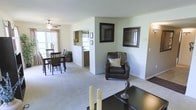 This single-floor residence showcases an open-concept floor plan that seamlessly connects the main living areas, creating a cohesive and functional flow throughout the space. The living area is anchored by plush wall-to-wall carpeting in a neutral tone, providing a warm and inviting foundation. A dark leather armchair, accented with a cream throw pillow, sits beneath a pair of framed decorative wall art pieces, adding character to the space.

A partial dividing wall separates the living area from the adjoining dining and kitchen zones without fully closing off the open feel. The dining area accommodates a dark wood table and matching chairs, positioned near a window that introduces natural light. A ceiling fan overhead serves both aesthetic and functional purposes in this area.

The kitchen is partially visible beyond the dividing wall, featuring a raised breakfast bar that connects to the dining space. Light-colored cabinetry and additional windows allow natural light to filter through, further brightening the interior. The overall color palette — soft greens, whites, and warm neutrals — lends the space a calm, understated ambiance suited to everyday living.