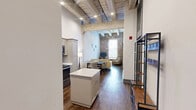 Located at 320 Andrew Higgins Drive in New Orleans' historic Warehouse District, this 2-bedroom, 2-bathroom unit blends industrial character with modern finishes. Soaring ceilings with exposed wooden beams and structural columns set a dramatic tone throughout, while rich hardwood-style flooring runs continuously across the entire single-level layout.

The open kitchen and living area anchors the space, featuring gray shaker cabinetry, white quartz countertops, a full suite of stainless steel appliances, a subway tile backsplash, and a central island with barstool seating. The adjacent living room is a standout, showcasing a full exposed brick accent wall and tall windows that flood the room with natural light — a nod to the building's historic industrial roots.

The primary bedroom offers generous proportions with soaring ceilings and direct access to a private en-suite bathroom, which includes a tub-shower combination, gray vanity, and wood-look tile flooring. The second bedroom is well-sized and served by a second bathroom featuring a walk-in shower. A hallway connects the private areas of the unit efficiently.

Throughout, the design balances the building's original architectural heritage — exposed brick, heavy timber, and raw structural elements — with clean, contemporary updates, resulting in a space that is both characterful and functional.