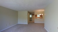 This two-floor residential space features an open-concept layout that connects the living area to the kitchen, creating a functional and cohesive flow. The living area is finished with wall-to-wall carpeting in a neutral tone, contributing to a warm and understated ambiance. Walls are painted in a soft, light beige, lending the space a clean and airy feel.

The kitchen is accessible via a pass-through counter that opens directly to the living area, allowing for easy interaction between the two spaces. Warm wood-toned cabinetry lines the kitchen walls, complemented by a white refrigerator and a sink positioned beneath a window that brings in natural light. A chandelier with multiple light fixtures hangs above the pass-through counter area, serving as a subtle decorative focal point.

A second window is visible toward the rear of the space, further enhancing natural light throughout. The transition between the carpeted living area and the kitchen's flooring is clearly defined. The overall design is practical and straightforward, with a neutral palette that provides a versatile foundation for a variety of interior styles.