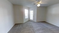 This single-floor residential space offers a clean and functional layout designed for comfortable everyday living. The main living area is finished with wall-to-wall carpet in a neutral gray tone, providing a soft underfoot feel throughout the space. Light-colored walls contribute to an open and airy atmosphere, while the textured ceiling adds a classic touch overhead. A ceiling fan with an integrated light fixture is centrally positioned, offering both air circulation and ambient lighting to the room. Natural light flows generously into the space through a pair of windows, including one situated in the corner that allows light from two directions, brightening the interior throughout the day. A paneled exterior door featuring a glass insert provides direct outdoor access and further supplements the natural light in the room. The door is complemented by a standard door frame and hardware, adding to the practical design of the space. Neutral wall tones and simple finishes give the space a versatile, move-in-ready quality that accommodates a variety of furnishing and décor styles. Overall, the space presents a straightforward and well-lit residential environment suited for single-level living.