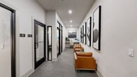 Located at 5000 Collina Ter in Clermont, FL, this single-floor office suite offers a well-appointed and efficiently designed professional environment. The layout comprises four distinct areas that flow cohesively, each finished with a consistent modern aesthetic defined by crisp white walls, neutral gray carpeting, and bold black-framed glass doors.

The reception corridor serves as a welcoming entry point, lined with a curated gallery of large-scale black-and-white framed artwork. Two cognac leather armchairs provide comfortable seating alongside a compact workstation, creating a polished and functional waiting area. Recessed lighting throughout ensures a bright, clean atmosphere.

Off the main corridor, dedicated cell rooms enclosed by sleek black-framed glass doors offer private spaces for focused work or calls, maintaining transparency while preserving quiet.

The conference room is a standout feature, centered around a substantial dark wood table surrounded by cushioned upholstered chairs. An elegant multi-arm chandelier with amber glass shades anchors the room, complemented by textured wall tile, three dimensional sculptural wall art panels, and a wall-mounted display screen — together creating a refined setting ideal for meetings and presentations.

Overall, this office suite balances contemporary design with practical functionality, delivering a professional and visually engaging workspace.