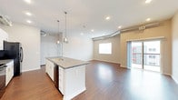 This single-floor unit showcases a contemporary design highlighted by an open-concept layout that seamlessly connects the kitchen, dining, and living areas. Rich, dark hardwood-style flooring runs throughout the main living spaces, complementing the neutral gray walls and white trim for a clean, modern aesthetic. Exposed metallic ductwork adds an industrial character overhead, tracing through the kitchen and living areas.

The kitchen is a standout feature, centered around a generous island with a double sink, gray stone-look countertops, and pendant lighting above. White cabinetry with brushed hardware lines the walls, paired with black appliances including a range, microwave, and side-by-side refrigerator. A dedicated in-unit laundry area accommodates a full-sized washer and dryer with overhead wire shelving.

The living area is bright and open, featuring recessed lighting and access to a private outdoor space through sliding glass doors. Bedrooms are carpeted for comfort, each receiving natural light through large windows with exterior views. The bathroom features a full tub-shower combination with white surround, while an elongated vanity with gray countertops and white cabinetry provides ample storage. A walk-in closet connects conveniently to the bathroom.