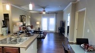 This single-story residence showcases an open-concept layout that seamlessly connects the kitchen and living areas, creating a cohesive and functional living environment. The rich, dark hardwood flooring runs throughout, providing a warm and polished foundation that ties the space together.

The kitchen is positioned at the front of the open layout, featuring granite-style countertops, a built-in sink, and a raised breakfast bar that overlooks the main living area. A pendant light fixture adds a modern touch directly above the counter space.

The living area is generously proportioned, accommodating a comfortable seating arrangement with a sofa, accent chairs, and a circular coffee table. Crown molding lines the perimeter of the ceiling, lending an elevated architectural detail to the space. A ceiling fan with integrated lighting sits centrally overhead, offering both airflow and ambient illumination.

Natural light pours in through a set of French doors dressed with white curtains and blinds, which also provide access to an outdoor area. A media console with a wall-mounted television anchors one side of the room, while a doorway beyond hints at additional rooms within the residence.