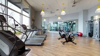 This fitness center is a thoughtfully designed, single-level facility that offers a comprehensive workout environment. The space is immediately welcoming, with an abundance of natural light streaming in through expansive floor-to-ceiling windows along one wall, creating a bright and energizing atmosphere.

The main cardio area features a row of Matrix treadmills aligned to take full advantage of the natural light and outdoor views, providing an engaging experience for users during their workouts. Complementing the cardio zone, a selection of indoor cycling bikes is positioned nearby, offering a dedicated space for high-intensity cycling sessions.

A full-length mirrored wall defines the strength and functional training area, visually expanding the space while allowing users to monitor their form. The area is well-equipped with a variety of free weights and training accessories.

Overhead, distinctive yellow pendant lights add a pop of color and a modern design touch against the neutral drop ceiling, while a ceiling fan ensures comfortable air circulation throughout. The warm-toned hardwood-style flooring ties the entire space together, providing both aesthetic appeal and a durable, easy-to-maintain surface. Overall, the facility strikes a clean, contemporary look that is both functional and inviting.