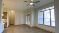 This single-story space features a clean, contemporary design centered around an open living area. The room is finished with warm-toned, wood-style hard flooring that extends throughout, providing a cohesive and low-maintenance foundation. Neutral white walls create a bright, versatile backdrop suitable for a variety of décor styles.

Natural light is a standout feature, with multiple windows fitted with horizontal blinds offering both illumination and privacy control. A set of glass-paneled doors—likely leading to a balcony or patio—further enhances the sense of openness and connectivity to the outdoors. A ceiling fan with an integrated light fixture is centrally mounted, contributing to both comfort and ambient lighting.

The layout appears efficient and well-proportioned, with a hallway visible in the background that suggests additional rooms beyond the main living space. Baseboards and door trim are finished in a complementary neutral tone, adding a subtle detail to the overall design. The space presents a move-in-ready aesthetic with a functional, modern feel throughout.