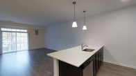 This single-story space showcases an open-concept design that integrates the kitchen and living areas into one cohesive environment. The kitchen island serves as a focal point, featuring a crisp white countertop surface and a stainless steel undermount sink, paired with dark cabinetry that creates a striking two-tone contrast. Two pendant lights hang above the island, providing focused task lighting while adding a contemporary design element to the space.

The dark hardwood-style flooring runs throughout, grounding the space with warmth and visual continuity. Neutral light gray walls contribute to a clean, modern aesthetic and allow for versatile decorating options. The open living area extends beyond the kitchen, offering generous room for furnishings and everyday use.

Practical details include a visible HVAC vent near the floor, ensuring climate control throughout the space. The layout promotes an easy flow between functional zones, making it well-suited for both daily living and entertaining. Overall, the space presents a modern, move-in-ready environment defined by clean lines and a cohesive neutral palette.
