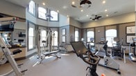 This fitness center is thoughtfully designed to support a comprehensive workout experience within an elegantly appointed space. The main exercise area impresses with dramatically high ceilings and a generous arrangement of windows at multiple levels, flooding the room with natural light. Decorative valances frame the windows, adding a refined aesthetic to what is typically a purely functional space. Ceiling fans provide continuous airflow throughout.

The cardio zone is well-stocked with a row of True-brand treadmills and elliptical machines, each equipped with individual display screens and positioned to face the windows. A cable strength machine with an adjustable bench anchors the free weights area, which also includes a dumbbell rack. A Peloton stationary bike and a recumbent cycle round out the cardio options. Wall-mounted mirrors expand the sense of space and support proper form during workouts.

A separate yoga and group fitness studio is enclosed by full-height glass sliding doors, featuring a dark hardwood floor, mirrored walls, a wall-mounted television, and yoga-themed decor. A water station and mounted flat-screen TV serve the main floor. The neutral gray and taupe color palette, recessed lighting, and polished finishes give the entire facility a modern, upscale character.