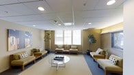 This single-floor space functions as a well-appointed waiting or reception area, designed to provide a comfortable and professional environment. The room features a drop ceiling with recessed lighting and a grid tile system, ensuring even, ambient illumination throughout the space. Warm beige and tan wall tones establish a neutral, calming palette complemented by blue-gray carpeting that anchors the seating arrangement.

Seating consists of several light-upholstered benches and chairs arranged around the perimeter and central area, accented with patterned throw pillows for added comfort and visual interest. A round glass coffee table with a metal frame serves as the centerpiece, topped with reading materials for guests. Horizontal blinds on the windows along the back wall allow for adjustable natural light.

Tall potted indoor trees positioned throughout the room add a touch of natural warmth and contribute to the welcoming ambiance. A framed artwork piece on the left wall adds a subtle decorative element. Overall, the space is thoughtfully arranged to balance functionality and comfort, making it well-suited for a professional or medical office setting.