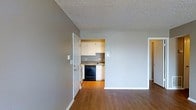 This single-floor space offers a straightforward and functional layout, with the main living area flowing directly into the kitchen. The living area is finished with warm hardwood-style flooring that extends throughout, providing a cohesive look across the space. Neutral grey walls give the interior a clean, understated appearance, while white baseboards and door trim add a crisp contrast.

The kitchen is accessible through an open pass-through from the main living area and is outfitted with white upper and lower cabinetry offering ample storage. A black dishwasher and range are integrated into the layout, complemented by a countertop workspace and a sink positioned along the back wall. Light grey tile backsplash ties the kitchen's finishes together neatly.

The textured popcorn ceiling is consistent throughout both spaces, and the layout benefits from a practical doorway configuration that allows for easy movement between rooms. Overall, the space presents a functional, no-frills interior suitable for everyday living, with durable finishes and a neutral palette that provides a versatile backdrop for personalization.