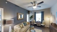 This thoughtfully designed single-floor space makes a strong first impression with its bold slate-blue accent wall, which serves as a striking backdrop in the main living area. The living room is anchored by a plush, neutral-toned sofa accented with patterned throw pillows in gold, teal, and green hues, complementing the overall color palette beautifully. A glass-top coffee table with ornate gold detailing adds a touch of glamour, while a decorative sunburst mirror mounted on the accent wall enhances the room's visual depth. Overhead, a classic five-blade ceiling fan with an integrated light fixture provides both comfort and ambient lighting. Dark hardwood-style flooring runs throughout, grounding the space with warmth and contrast against the lighter furnishings. Natural light pours in through multiple large windows dressed with teal curtain panels, creating an airy and inviting atmosphere. A lush indoor plant adds a refreshing organic element near the entryway. The dining area, visible from the living room, features an elegant chair arrangement, reinforcing the cohesive and curated aesthetic of the space. Overall, the design balances modern sophistication with comfortable, livable style.