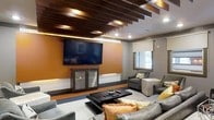 This single-floor space showcases a thoughtfully designed lounge area that blends contemporary aesthetics with comfortable functionality. The focal point of the room is a large wall-mounted flat-screen television set against a rich, warm amber accent wall, creating a bold visual contrast against the surrounding neutral gray walls. Below the screen, a low-profile media console with glass-front cabinets provides storage while maintaining a sleek, uncluttered appearance. A subtle raised platform adds architectural dimension to the entertainment area.

One of the most distinctive features of the space is the ceiling design, where dark-stained wooden beams are arranged in a parallel pattern across a white ceiling, complemented by recessed lighting that highlights the beams and adds warmth to the room. The seating arrangement includes plush gray sofas and armchairs styled with accent pillows in gold and green tones, centered around a dark rectangular coffee table. Natural light filters in through a window fitted with a neutral roller shade. The overall ambiance is modern and inviting, balancing bold design choices with a relaxed, lounge-like atmosphere.
