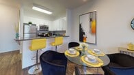 This two-story residence offers a thoughtfully designed layout that balances everyday functionality with modern style. The main level opens into a seamless kitchen and living area, where rich hardwood-style flooring flows throughout. The kitchen features white cabinetry, dark countertops, stainless steel appliances including a dishwasher and refrigerator, and a peninsula with bar seating accented by bold yellow chairs. Track lighting overhead adds a contemporary touch. Adjacent to the kitchen, the dining area transitions naturally into a spacious living room bathed in natural light from large vertical-blind windows. A sliding glass door connects the living area to a private outdoor patio. A carpeted staircase with a warm wood handrail leads to the upper level, where the bedrooms are located. Each bedroom features plush carpet, generous closet space, and large windows that invite natural light. The bathrooms are clean and well-appointed, with white shaker-style vanities, dark countertops, and modern fixtures. One bathroom includes a walk-in closet nearby. The entry area on the main floor includes a decorative console with a full-length mirror, offering a welcoming first impression. Throughout the residence, a neutral color palette creates a calm, cohesive ambiance.