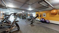 This fitness facility offers a comprehensive workout environment designed to accommodate a wide variety of training styles and fitness goals. The space features an industrial aesthetic, with exposed ceiling infrastructure including ductwork and piping that lend the gym a raw, urban character. Durable rubber flooring throughout provides both safety and comfort underfoot.

The gym is generously outfitted with an impressive array of equipment. Cardio machines, including an elliptical and treadmill, are positioned toward the front of the space, while a robust selection of strength-training machines occupies the central floor area. A dedicated free weights section includes a full dumbbell rack, and functional training equipment such as an inversion table, medicine balls, and stability balls round out the offering.

A vibrant yellow accent wall adds a burst of energy to one side of the facility, complemented by motivational artwork. Full-length mirrors are strategically placed to maximize the sense of space and assist with form during workouts. Overhead lighting ensures the entire area is well-lit, creating a focused and energizing atmosphere. This facility is thoughtfully arranged to make efficient use of its footprint while keeping all equipment highly accessible.