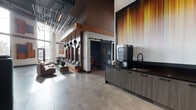 Located at 1101 N Main St in Los Angeles, CA, this contemporary single-floor lobby space makes a striking first impression with its soaring double-height ceilings and an industrial-chic aesthetic that seamlessly blends warm natural wood with dark metal accents. Large-format stone tile flooring runs throughout, grounding the space with a sense of solidity and polish.

The lounge area serves as the social heart of the lobby, featuring a built-in wood-paneled banquette with deep charcoal cushions, accented by a series of bold circular artworks in vibrant amber, red, and blue tones. Pendant lights with matte black shades drop from a richly detailed wood soffit, casting a warm, inviting glow over the seating arrangement. Complementary lounge chairs and low coffee tables complete the gathering space.

A dedicated media nook offers a high-back dark sofa positioned beside floor-to-ceiling windows that draw in natural light and views of the surrounding streetscape, paired with a wall-mounted display screen for entertainment or information.

The kitchenette area features sleek dark cabinetry with black hardware, a built-in sink, and a beverage station, providing practical amenities within the common space. A large-scale gradient mural in warm amber and gold tones anchors one wall, adding a dramatic artistic focal point. A sliding barn door leads to an additional private area, rounding out this thoughtfully designed, multifunctional lobby.