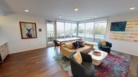 Located at 2390 SE Hawthorne Blvd in Portland's vibrant Hawthorne neighborhood, this contemporary single-floor space offers a thoughtfully designed interior with a clean, modern aesthetic. The main living area is a standout, anchored by rich hardwood flooring and an expansive wall of floor-to-ceiling windows and sliding glass doors that flood the room with natural light and open onto a private outdoor balcony with treetop views. The open layout comfortably accommodates both a lounge seating arrangement — featuring a sofa, swivel chairs, a coffee table, and a wall-mounted flat-screen TV atop a natural wood media console — and a separate dining area with a wooden table. Bold artwork, including a colorful mixed-media flag piece, adds a curated, creative character to the white walls. Recessed lighting throughout ensures a well-lit environment at any time of day. A compact kitchenette offers light wood cabinetry, quartz countertops, a double sink, microwave, beverage refrigerator, and open floating shelves set against a herringbone white tile backseat with warm under-shelf lighting. A clean, minimalist hallway with hardwood flooring leads to the entryway, completing this polished and functional space.