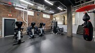 Located at 200 Rhode Island Ave NE in Washington, DC, this fitness center combines industrial aesthetics with a well-equipped workout environment. The space occupies a single open floor and is characterized by its striking exposed brick walls, dark ceiling with visible ductwork and piping, and robust concrete support columns — all contributing to a raw, urban atmosphere. Durable rubber flooring spans the entire area, providing a safe and functional surface for a wide range of exercises. The cardio zone features multiple True treadmills and elliptical machines, complemented by a wall-mounted flat-screen television for entertainment during workouts. A dedicated strength training area includes a multi-station cable and weight machine, along with free weights and battle ropes for functional training. A UFC-branded heavy punching bag and speed bag round out the boxing station, adding a combat fitness dimension to the space. Large wall mirrors along one section visually expand the room and allow users to monitor their form. Natural light filters in through several ground-level windows, while overhead fluorescent strip lighting ensures consistent illumination throughout. The open layout allows for versatile use of the space, accommodating both individual training sessions and group workouts with ease.