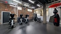 Located at 200 Rhode Island Ave NE in Washington, DC, this fitness center combines industrial aesthetics with a well-equipped workout environment. The space occupies a single open floor and is characterized by its striking exposed brick walls, dark ceiling with visible ductwork and piping, and robust concrete support columns — all contributing to a raw, urban atmosphere. Durable rubber flooring spans the entire area, providing a safe and functional surface for a wide range of exercises. The cardio zone features multiple True treadmills and elliptical machines, complemented by a wall-mounted flat-screen television for entertainment during workouts. A dedicated strength training area includes a multi-station cable and weight machine, along with free weights and battle ropes for functional training. A UFC-branded heavy punching bag and speed bag round out the boxing station, adding a combat fitness dimension to the space. Large wall mirrors along one section visually expand the room and allow users to monitor their form. Natural light filters in through several ground-level windows, while overhead fluorescent strip lighting ensures consistent illumination throughout. The open layout allows for versatile use of the space, accommodating both individual training sessions and group workouts with ease.