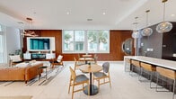 Located in Miami, FL, this thoughtfully designed space blends mid-century modern aesthetics with contemporary finishes across a single well-appointed floor. The open-concept layout seamlessly connects the kitchen, dining, and lounge areas, creating an inviting and cohesive environment throughout.

The kitchen is a standout feature, anchored by an expansive white waterfall island with seating for multiple guests. Dark espresso cabinetry lines the walls, complemented by stainless steel appliances and a series of globe pendant lights with brass fixtures that cast warm, ambient light across the space.

The lounge area exudes comfort and style, with rich wood slatted wall paneling serving as a dramatic backdrop for a wall-mounted flat-screen TV and a sleek built-in fireplace below. Tufted leather seating, mid-century accent chairs, and a circular white coffee table are anchored by a patterned area rug. A sculptural ring chandelier overhead adds an artistic focal point. Floor-to-ceiling windows flood the space with abundant natural light and provide views of the surrounding greenery.

The dining area features round tables with mixed seating arrangements, set against warm wood-paneled walls and large windows that connect the interior to the outdoors. A balcony extends the living space further, offering an outdoor retreat. Large-format porcelain tile flooring ties the entire space together with a clean, polished finish.