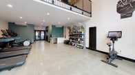 This fitness center offers a thoughtfully designed space for a comprehensive workout experience. The layout is open and spacious, with polished concrete floors that give the space a clean, contemporary aesthetic. Large floor-to-ceiling windows and glass double doors along one wall flood the interior with natural light while providing views of an outdoor courtyard area, creating an inviting atmosphere.

The cardio section is well-appointed, featuring multiple treadmills and elliptical machines, each equipped with individual digital screens for entertainment and performance tracking. Stationary and cycling bikes, including a Peloton, are also available for low-impact cardio training. A dedicated functional training zone includes TRX suspension straps, medicine balls, plyo boxes, and additional resistance equipment, all neatly organized on a multi-tiered storage rack.

A built-in countertop station with an under-counter refrigerator provides a convenient spot for hydration and post-workout recovery. The space is climate-controlled with ceiling-mounted HVAC units. An upper mezzanine railing is visible overhead, adding architectural dimension. Teal accent walls throughout add a modern, energetic touch, complemented by subtle wall art that enhances the overall motivating ambiance.