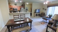This single-floor space offers an open-concept layout that seamlessly connects the kitchen, dining, and living areas, creating a cohesive and functional environment. The kitchen is well-appointed with stainless steel appliances, including a microwave, oven, and refrigerator, complemented by warm wood cabinetry and light-toned countertops. A peninsula counter provides additional workspace and serves as a natural divider between the kitchen and the rest of the living area. The dining area features a rich, dark wood farmhouse-style table paired with matching chairs and a bench, lending a rustic yet homey character to the space. Crown molding along the ceiling adds a refined architectural detail throughout. The living room is carpeted and comfortably arranged with seating and a media console, creating an inviting area for relaxation and entertainment. A ceiling fan provides airflow, while wall-mounted accent lighting adds warmth to the ambiance. The neutral color palette of soft beige tones throughout ties all the areas together, resulting in a cohesive and welcoming atmosphere that feels both practical and lived-in.