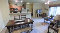 This single-floor space offers an open-concept layout that seamlessly connects the kitchen, dining, and living areas, creating a cohesive and functional environment. The kitchen is well-appointed with stainless steel appliances, including a microwave, oven, and refrigerator, complemented by warm wood cabinetry and light-toned countertops. A peninsula counter provides additional workspace and serves as a natural divider between the kitchen and the rest of the living area. The dining area features a rich, dark wood farmhouse-style table paired with matching chairs and a bench, lending a rustic yet homey character to the space. Crown molding along the ceiling adds a refined architectural detail throughout. The living room is carpeted and comfortably arranged with seating and a media console, creating an inviting area for relaxation and entertainment. A ceiling fan provides airflow, while wall-mounted accent lighting adds warmth to the ambiance. The neutral color palette of soft beige tones throughout ties all the areas together, resulting in a cohesive and welcoming atmosphere that feels both practical and lived-in.