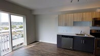 This single-floor space showcases a clean, modern design with an open-concept layout that seamlessly connects the living area and kitchen. The flooring throughout consists of rich, dark hardwood-style planks that add warmth and contrast to the otherwise light and neutral palette of the space.

The kitchen is outfitted with two-toned cabinetry, featuring dark lower cabinets paired with light wood-grain upper cabinets with vertical striping, creating a striking visual contrast. The countertops are bright white, complemented by a white subway tile backsplash that reinforces the contemporary aesthetic. A stainless steel dishwasher is integrated into the lower cabinetry. A track lighting fixture is mounted on the ceiling above the kitchen, providing focused illumination.

The living area is open and airy, with neutral white walls and smooth ceilings that enhance the sense of space. A large floor-to-ceiling window and sliding glass door along one wall flood the interior with abundant natural light and offer views to the outdoors. The overall ambiance is minimalist, bright, and move-in ready, suited to a modern lifestyle.