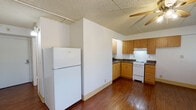 This single-floor unit is laid out efficiently, offering a functional and straightforward living environment. The main living area is open and well-lit, benefiting from a wide window fitted with blinds that allows natural light to fill the space. A ceiling fan with a light fixture is centrally installed, providing both air circulation and ambient lighting. The floors throughout the unit are finished with rich, dark wood-style planking, adding warmth to the otherwise neutral white walls.

The kitchen occupies a dedicated L-shaped area, outfitted with warm-toned wooden cabinetry extending to the upper and lower walls, dark laminate countertops, a stainless steel sink, a white electric range, and a full-size white refrigerator. The textured drop ceiling is consistent throughout the unit, lending a cohesive look to the interior.

A wall-mounted sconce light fixture illuminates the entryway corridor, which provides access to a closet with built-in shelving. The bathroom is clean and functional, featuring white tile walls surrounding a full bathtub with shower, a toilet, and an oak-finished vanity with a single sink and medicine cabinet mirror above. The floor is finished with small mosaic tiles.