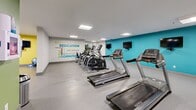 Located at 1020 Paisley Rd in Guelph, Ontario, this fitness centre offers a comprehensive, single-floor workout facility designed to support a wide range of exercise routines. The open-plan layout provides ample space for movement, with a durable speckled flooring surface underfoot and bright, recessed panel lighting overhead that ensures the entire area is well-illuminated.

The cardio zone is prominently featured, housing a row of Matrix-brand treadmills and elliptical trainers positioned alongside a teal accent wall. Wall-mounted flat-screen televisions are conveniently placed for viewing during workouts. The strength training area is anchored by a full-length mirror wall that enhances the sense of openness, and includes a dumbbell rack, weight bench, and multi-function strength machines. Stability balls and additional accessories are neatly stored nearby.

The facility is further distinguished by vibrant accent walls in teal, green, and yellow, along with a bold motivational typography mural featuring words such as "Dedication," "Commitment," and "Aim Higher." Safety is addressed through a clearly mounted AED (defibrillator) station. Decorative touches, including framed artwork and a tall ornamental plant, contribute to an energetic yet welcoming atmosphere. Overall, the space is thoughtfully organized to accommodate both cardio and strength training needs efficiently.