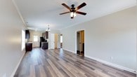This single-story cottage at The Cottages offers a thoughtfully designed open-concept interior that blends comfort and modern functionality. The main living area is characterized by rich, wood-toned luxury vinyl plank flooring that extends throughout, creating a warm and cohesive aesthetic. Crown molding lines the ceiling, adding a refined architectural detail, while a ceiling fan with a light fixture provides both comfort and ambient lighting.

The open layout allows the living space to flow naturally into the kitchen area, which features dark cabinetry paired with stainless steel appliances, including a range and microwave. A pendant chandelier hangs above the kitchen transition zone, adding a decorative touch. Natural light filters in through multiple windows, keeping the interior bright and inviting.

Neutral gray-toned walls complement the flooring and trim throughout, creating a clean, versatile palette. Doorways visible from the main living space lead to additional rooms, suggesting a practical and well-organized floor plan. The overall ambiance is bright, modern, and move-in ready, with finishes that balance style and durability throughout.