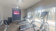 Located at 1800 Arboretum Cir in Vestavia Hills, Alabama, this single-floor fitness center is designed to support a well-rounded workout experience. The space is finished with neutral gray walls and durable multi-tone carpet tile flooring, creating a clean, modern aesthetic throughout. Large floor-to-ceiling windows line one side of the room, flooding the interior with abundant natural light and offering views of the surrounding outdoor greenery — an energizing backdrop for any workout.

The cardio area is well-equipped with multiple Matrix treadmills and elliptical machines positioned to take full advantage of the window views. A wall-mounted Vizio 4K HDR Smart TV is featured on a slate-blue accent wall, providing entertainment during cardio sessions. A ceiling fan overhead helps maintain comfortable airflow throughout the space.

The strength and functional training area includes a rack of rubber hex dumbbells in a range of weights, a flat weight bench, a cable machine, and a spin bike, offering versatile options for both strength training and cycling. Exercise balls in assorted sizes are neatly stored on a rack near the rear of the room, supporting stretching and balance training. A glass-paned door at the back provides additional access to the exterior, further enhancing natural light and ventilation. The overall ambiance is bright, functional, and welcoming.