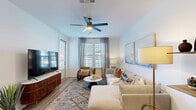 This thoughtfully designed one-bedroom, one-bathroom residence offers an efficient and well-organized single-floor layout. The open-concept living and kitchen area forms the heart of the unit, where light wood-tone flooring, white walls, and abundant natural light from plantation-shuttered windows create a bright, welcoming atmosphere. A ceiling fan with integrated lighting anchors the living space, which comfortably accommodates a sectional seating arrangement.

The kitchen is a standout feature, showcasing white shaker-style cabinetry, granite countertops, a large center island with a built-in sink and seating, stainless steel appliances, a subway tile backsplash, and pendant lighting. The layout provides ample storage and prep space.

The carpeted primary bedroom offers comfortable proportions, natural light through shuttered windows, and a ceiling fan overhead. A dedicated closet provides storage convenience.

The bathroom mirrors the kitchen's design sensibility with white cabinetry, granite countertops, a bold black-framed vanity mirror, and light wood-tone flooring, creating a clean, cohesive aesthetic throughout the entire residence.