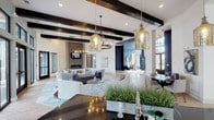 This sophisticated single-floor community space offers an open-concept design that seamlessly blends comfort and modern elegance. The main lounge area is anchored by a sleek linear fireplace set against a stacked-stone surround, accompanied by plush seating arrangements and a wall-mounted television — an inviting setting for gathering and relaxation. Striking exposed dark wood ceiling beams contrast beautifully against crisp white walls, while an array of oversized glass pendant lights and a circular chandelier lend warmth and character throughout. A dark granite-topped bar counter runs along one side of the space, accented by live plant arrangements that add a natural touch. Large floor-to-ceiling black-framed windows and glass doors flood the interior with natural light and offer views of the outdoor surroundings. Adjacent to the lounge, a dedicated game room features a full-size billiards table set atop a patterned area rug, with additional pendant lighting and bar-height seating nearby. A refreshment station equipped with a coffee machine and under-counter refrigeration provides convenient amenities for guests. Light wood-tone flooring flows throughout, tying the various areas together into a cohesive, well-appointed space designed for community engagement and leisure.