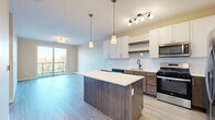 Located at 101 S Main St in Lombard, Illinois, this single-floor residence offers a thoughtfully designed open-concept layout with modern finishes throughout. The kitchen serves as a central feature, showcasing a large island with a quartz countertop and warm wood-toned cabinetry complemented by crisp white upper cabinets. Stainless steel appliances, including a refrigerator, gas range, microwave, and dishwasher, are paired with pendant lighting and a track lighting system, creating a well-lit, functional workspace that flows seamlessly into the living area. Light-toned hardwood-style flooring runs throughout the main living spaces, enhancing the sense of openness and continuity. The living area benefits from abundant natural light through a sliding glass door that opens to a private balcony with outdoor views of the surrounding Lombard streetscape and greenery. The bedroom features plush carpeting, large windows with exterior views, and a built-in closet with shelving for organized storage. The bathroom is appointed with a sleek frameless sliding glass shower enclosure and matte black hardware accents. A dedicated in-unit laundry area houses a full-sized Whirlpool washer and dryer. The overall design aesthetic is clean, contemporary, and functional, making this a well-appointed urban residence.