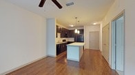 This single-story residence at Capital Place At Southwood showcases an open-concept layout that seamlessly connects the living and kitchen areas, creating a spacious and functional living environment. Rich, dark hardwood flooring runs throughout, adding warmth and elegance to the space.

The kitchen is a standout feature, appointed with deep espresso-toned cabinetry that extends to the upper walls, providing generous storage. Black appliances complement the dark cabinetry, while a central island with a light countertop offers additional workspace and casual dining potential. Pendant lighting hangs above the island, casting a warm, inviting glow. A decorative tile backsplash adds a subtle design accent to the cooking area.

The adjoining living area benefits from the open layout, enhanced by a ceiling fan that promotes air circulation. Neutral walls throughout provide a clean, versatile backdrop suitable for a variety of décor styles. A hallway visible in the background leads to additional rooms, hinting at a well-organized floor plan. The unit's entry door is conveniently accessible, rounding out this thoughtfully designed single-floor residence.
