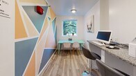 Located at 360 Main St in Hartford, Connecticut, this compact yet thoughtfully designed business center offers a highly functional and visually engaging workspace. The room is defined by its striking geometric mural wall, featuring a dynamic arrangement of peach, gold, sky blue, and teal triangular shapes that instantly energize the space and serve as a bold artistic focal point.

The flooring throughout is light-toned wood-look vinyl plank, lending warmth and a clean, modern aesthetic. A granite-topped counter runs along one wall, outfitted with computer workstations and bar-height seating — ideal for focused individual work or printing tasks. A second dedicated workspace features a teal table positioned directly beneath a bright window, which frames lush, mature tree canopy outside and floods the room with natural light.

Entry is through a solid wood door with a gold lever handle, opening into a welcoming corridor that connects to the broader building. The walls are finished in a neutral white, allowing the colorful mural to command attention without overwhelming the space. Overhead lighting and HVAC vents are cleanly integrated into the ceiling. The overall ambiance is lively and modern, balancing practical amenities with a creative, upbeat atmosphere well-suited for a shared community or business hub.