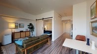 This well-designed single-floor unit combines contemporary style with functional living. The open-concept living area is anchored by rich hardwood-style flooring and bathed in natural light from expansive floor-to-ceiling windows that frame sweeping urban views. A leather sofa, curated artwork, and a compact dining area create a comfortable and stylish gathering space.

A standout architectural feature is the sliding wood barn door with black hardware, which elegantly separates the bedroom from the main living area. The bedroom is a calm retreat, furnished with a queen bed, warm bedside lighting, beach-themed artwork, and a built-in sliding-door closet for ample storage.

The galley-style kitchen runs efficiently along one wall, featuring stainless steel appliances, white cabinetry, open wooden shelving, a tile backsplash, and a clean countertop workspace — all flowing toward the unit's entry corridor.

The bathroom is finished in a clean, modern palette with wood-look flooring, a white vanity with a stone-toned countertop, an undermount sink, and a full soaking tub with shower. Throughout the unit, a consistent design language of neutral whites, warm wood tones, and modern fixtures creates a cohesive and inviting ambiance suited for comfortable everyday living.