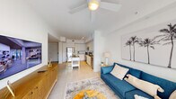 This single-floor unit showcases a clean, modern design defined by crisp white walls, large-format tile flooring, and an open layout that seamlessly connects the main living spaces. The living area is bright and airy, benefiting from natural light filtering through a window dressed with sheer draping. A ceiling fan overhead adds a casual, coastal feel, complemented by a large palm tree artwork that reinforces the relaxed aesthetic. A wall-mounted flat-screen TV and warm wood media console ground the space.

The kitchen is a standout feature, equipped with stainless steel appliances including a refrigerator, range, and built-in microwave. Tall white cabinetry extends to the ceiling, providing generous storage, while dark countertops offer a striking contrast. A central island with seating serves as both a practical workspace and casual dining area.

The bedroom is a serene retreat with tile flooring, a ceiling fan, and a window allowing natural light. Soft, neutral tones and layered textiles create a calm atmosphere.

The bathroom maintains the unit's contemporary palette with white cabinetry, a dark countertop vanity, large tile flooring, and a glass-enclosed walk-in shower, offering a functional and polished finish throughout.