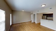 This single-floor space is designed with an open and functional layout that maximizes the sense of spaciousness. The main living area is finished with warm-toned hardwood-style flooring that extends throughout, complementing the neutral beige walls and white trim for a clean, cohesive aesthetic. Flush-mount ceiling light fixtures provide consistent overhead lighting, while HVAC vents integrated into the ceiling contribute to a comfortable interior environment.

The open floor plan seamlessly connects the living area to the kitchen, which is partially visible to the right and features a white peninsula counter and a stainless steel refrigerator, offering a practical layout for daily use. Toward the rear of the space, a white paneled door leads to an additional area, alongside a built-in shelving unit that provides organized storage. The white shelving adds a structural detail that enhances both utility and visual interest. The smooth white ceilings and understated color palette throughout create a calm, neutral ambiance well-suited to a variety of interior styles and furnishing arrangements.