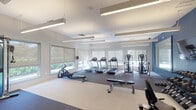 This fitness center is designed to support a comprehensive workout experience within a single, open-plan layout. The space is defined by its clean, contemporary aesthetic, featuring white walls accented by a bold blue feature wall that adds visual depth and energy to the environment. Suspended linear lighting fixtures illuminate the room evenly, complementing the abundant natural light that streams in through a series of large windows fitted with roller shades, offering views of surrounding greenery.

The cardio zone is well-appointed with multiple treadmills, elliptical cross-trainers, and a rowing machine, all arranged to take advantage of the window views. A dedicated strength training area features a dumbbell rack stocked with a full range of weights, a weight bench, and a cable machine system, providing options for both free-weight and machine-based resistance training. Stability balls and additional functional fitness equipment are also available for stretching and core work.

Large wall-mounted mirrors expand the sense of space and aid in proper form during exercise. A wall-mounted flat-screen television enhances the training environment. The entry area includes a small storage unit and posted fitness center policies, offering a tidy and organized welcome. The flooring transitions between cushioned rubber matting in the active zones and lighter surfaces near the perimeter, prioritizing both safety and comfort throughout.