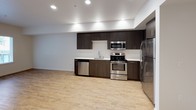 This single-floor unit offers a well-designed layout with modern finishes. The kitchen serves as a functional centerpiece, featuring dark espresso cabinetry extending to the ceiling, white countertops, and a full suite of stainless steel appliances including a refrigerator, electric range, microwave, and dishwasher. Light wood-style flooring runs throughout the main living and kitchen areas, complemented by recessed lighting that creates a bright, open atmosphere. A convenient in-unit stacked washer and dryer is tucked neatly into a dedicated alcove near the entryway, adding practical value to the space.

The bedroom provides a comfortable retreat with plush carpeting underfoot and a corner window configuration that invites natural light while offering views of the surrounding exterior. The neutral wall tones keep the room feeling calm and versatile.

The bathroom is finished with a dark wood-grain vanity topped with a white countertop and undermount sink, a large mirror with overhead lighting, open shelving for storage, and wood-look tile flooring. A tub-shower combination completes the space. Overall, the unit presents a clean, contemporary aesthetic with thoughtful attention to storage and functionality.