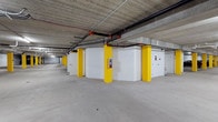 This single-level parking structure is defined by its functional, utilitarian design and generous open layout. Bold yellow-painted structural columns run the length of the facility, providing clear visual markers throughout the drive aisles while contributing a striking contrast against the white-painted concrete block walls. The exposed concrete ceiling overhead reveals an organized network of pipes and conduit, including a prominently routed fire suppression system with bright red fittings, underscoring the facility's commitment to safety compliance. The smooth, light grey concrete floor is clean and well-maintained, extending across a wide expanse that allows for comfortable vehicle navigation. White-painted masonry block enclosures are positioned throughout the interior, likely housing mechanical or utility systems, and are fitted with fire extinguisher stations for added safety. Access doors are evenly spaced along the perimeter walls, facilitating pedestrian movement throughout the structure. The overall ambiance is bright, open, and orderly, with the high ceiling clearance and unobstructed sightlines creating a sense of spaciousness well-suited to a high-capacity parking environment.